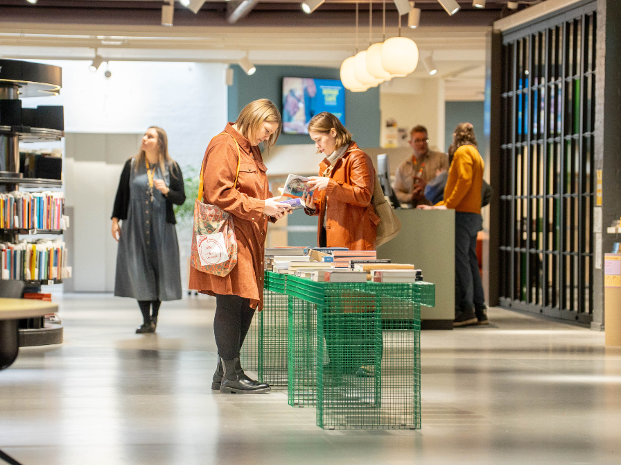 Billede af to biblioteksbrugere ved et grønt trådbord. De kigger på bøger. Bibliotekspersonale i baggrunden. 